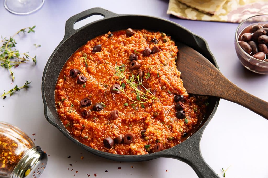 A cast iron skillet filled with a vibrant vegan puttanesca made with quinoa, tomatoes, capers, and sliced kalamata olives, shown with a wooden spatula and a sprig of fresh thyme
