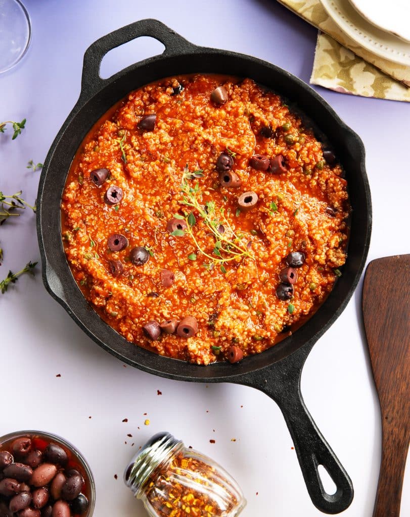 Overhead view of a rustic puttanesca sauce tossed with fluffy quinoa and topped with sliced black olives, tiny capers, and a fresh thyme sprig in a cast iron skillet.