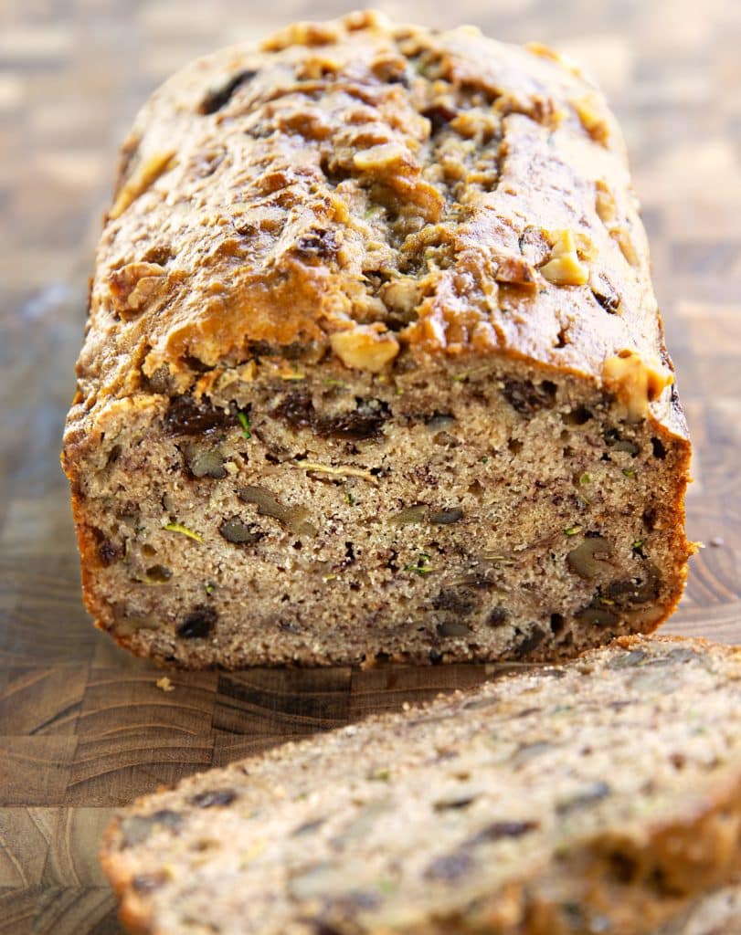 Close-up view of a sliced loaf of vegan zucchini banana bread, showing a thick cross-section filled with chopped walnuts and raisins under a textured, golden crust.