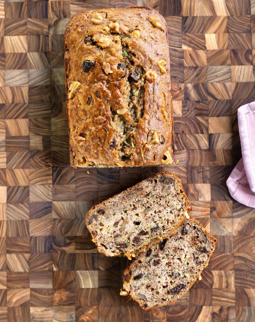 Overhead view of a golden-brown loaf of vegan zucchini banana bread on a wooden cutting board, with one slice cut and placed next to it.